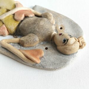 Figurka do zawieszenia w postaci Anioła, anioł dziewczyna, figurka przedstawiająca kobietę ze skrzydłami Anioła, kobieta ma blond włosy z kokiem na czubku głowy, ubrana jest w bluzkę z bufiastymi rękawami oraz szerokie, żółte spodnie kuloty, w ręku oraz w kieszeniach są serca.